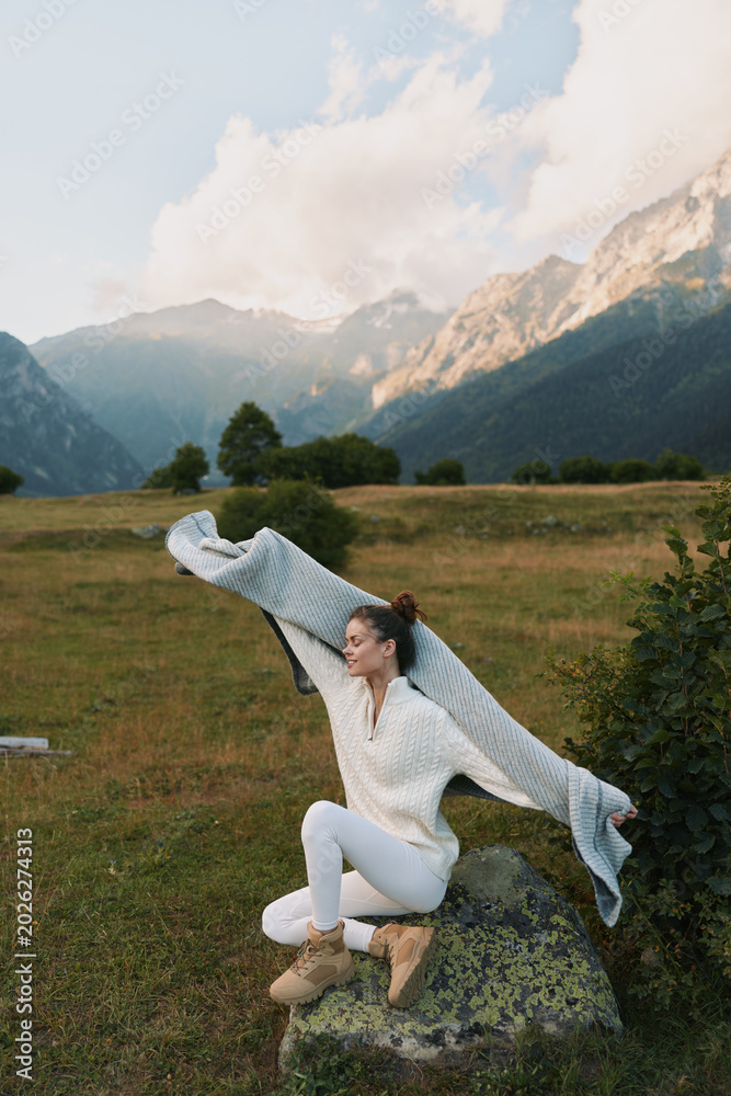Fototapeta premium A woman in a white sweater and boots leaps across a meadow, with mountains in the background, capturing a vibrant outdoor lifestyle moment in nature.