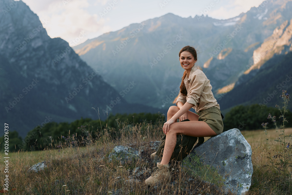 Fototapeta premium A young woman sits on a rock in a mountain meadow, smiling softly as she enjoys a peaceful, outdoor escape with rugged peaks in the distance.