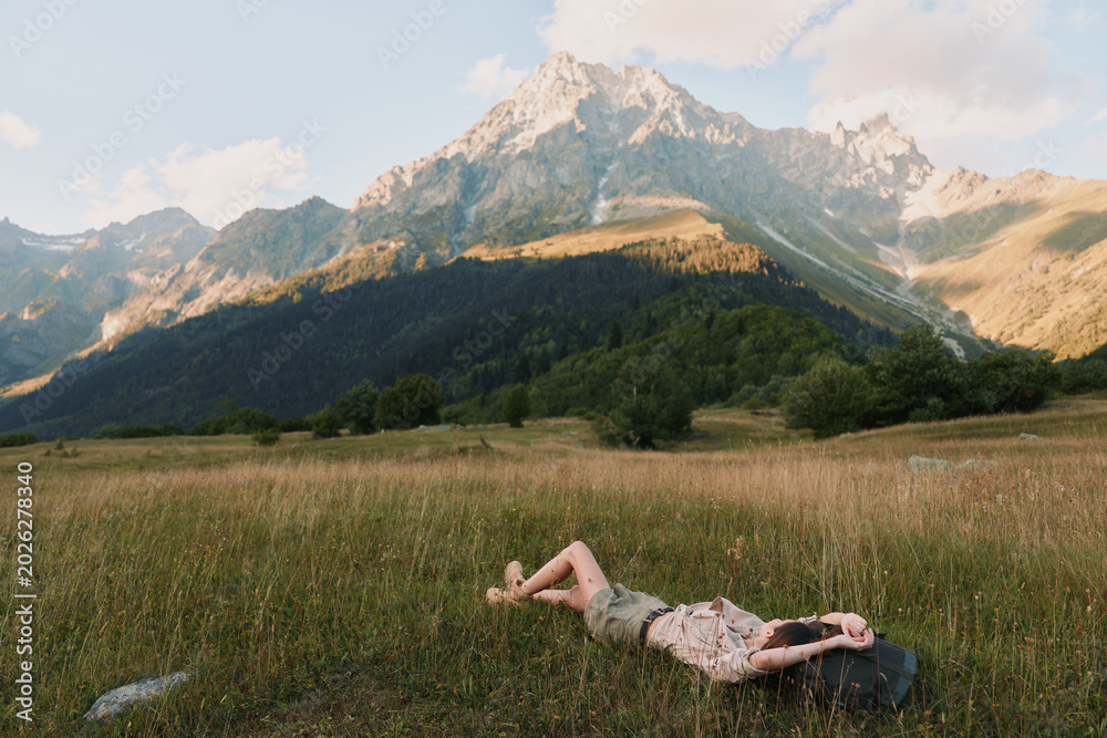Fototapeta premium A woman in casual clothes lies on grass in a mountain meadow, relaxing under a partly cloudy sky with tall peaks in the background, enjoying nature and lifestyle outdoors.