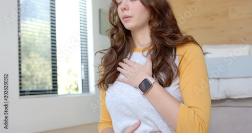 Adult woman placing hands on chest and belly, breathing to relax at bed wearing gray shirt