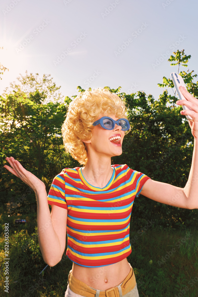 Fototapeta premium Young woman with curly blonde hair wearing a striped rainbow shirt and blue sunglasses poses for a joyful outdoor selfie in a sunlit park, the bright colors echo a retro vibe and a carefree outdoor