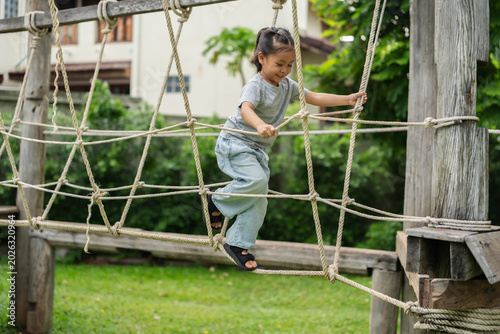preschool child girl playing and walking on wooden rope bridge at outdoor playground in park
