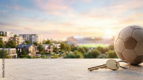 Real estate investment near sports stadium concept with soccer ball and house key on balcony overlooking residential area and arena at sunset
