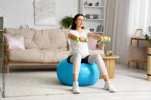 Beautiful young pregnant woman training with fitball and dumbbells at home