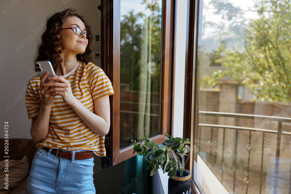 Fototapeta premium Young woman stands by an open window at home, gazing outside while holding a smartphone, wearing a striped yellow shirt and jeans, with a plant on the sill and bright natural light.