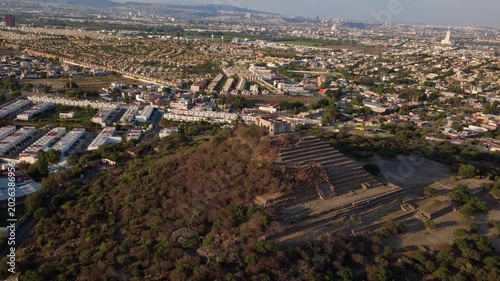 Drone tilt down shot over ancient pyramid ruins in El Cerrito Querétaro Mexico revealing archaeological structures surrounded by modern urban landscape at sunset