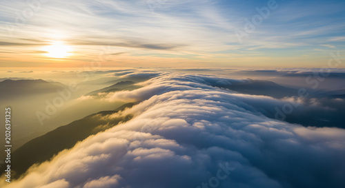 Beautiful aerial cloudscape above high mountain summit with thick soft clouds flowing gracefully between dramatic peaks, early morning sun casting glowing gold and pastel colors across the sky