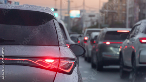 Behind a white car with its red brake lights illuminated during evening traffic with drop rain on car. Vehicles stretches ahead on a crowded city street as dusk begins to settle urban landscape.