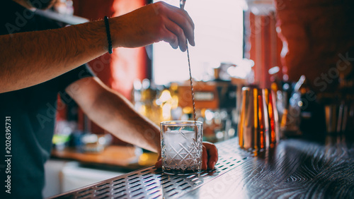 Cocktail making. Ice cube mixing. Unrecognizable barman hands stirring alcohol drink with professional spoon in transparent glass on bar counter.