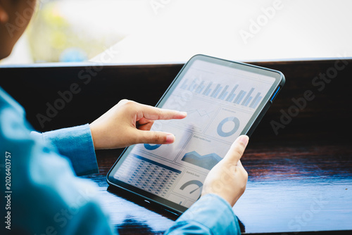 Close-up of person using tablet with business analytics charts at wooden table near window, blurred outdoor background, modern workspace, data dashboard on screen.