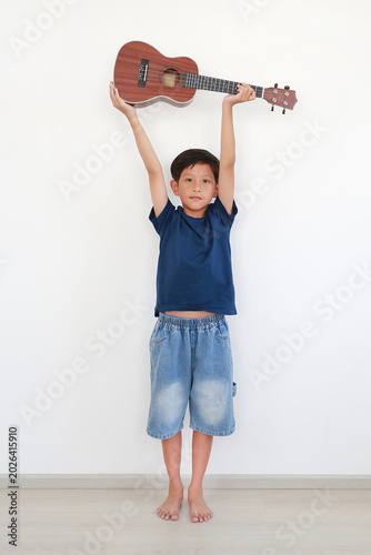 Portrait Asian boy standing and holding ukulele with both hands up high over head against white wall background.