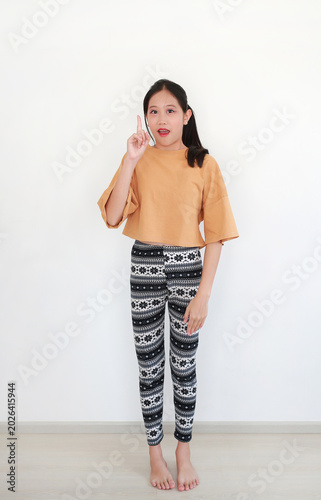 Smiling young Asian girl standing, pointing an index finger upward with a look of surprise on her face, in front of white wall.