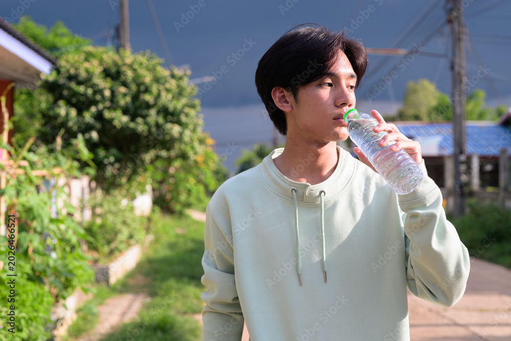 Fototapeta premium Young Thai man from Isan drinking bottled water looking away outdoors