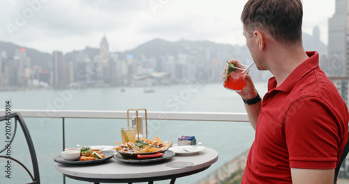 Man in red polo shirt drinking cocktail at rooftop bar facing Hong Kong skyline and Victoria Harbour. luxury lifestyle, travel destination, urban dining, vacation, metropolis, asian tourism.