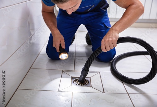 A worker kneels, shining a flashlight into a wet floor drain while using a hose to clear a blockage.