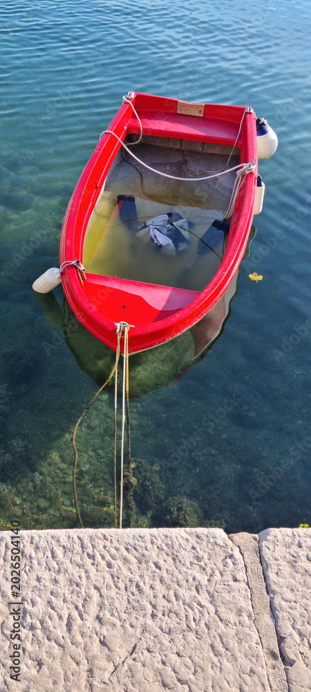Obraz premium High angle view of a small red plastic rowboat filled with water and tied to a stone pier in the clear sea.