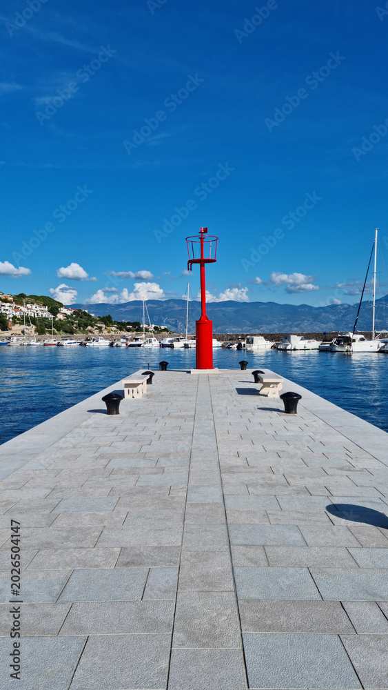 Obraz premium Symmetrical low angle view of a red harbor lighthouse at the end of a stone pier in Baska on Krk island Croatia.