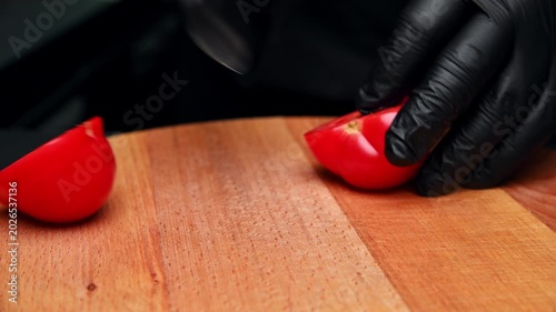Chef slices red tomato on wooden cutting board with black gloves in kitchen