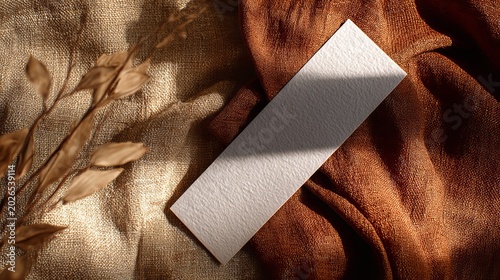A blank white card on a textured brown fabric background with dried plants