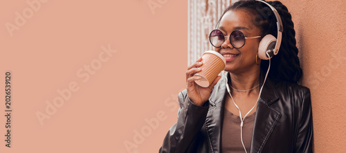 Fashionable black woman with headphones and a takeaway leaning against a pink wall, copy space