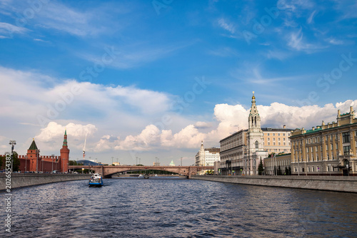 Bright sky over the Moscow river shows a boat passing by. The buildings along the river feature historical architecture. The scene captures an outdoor view of the city.