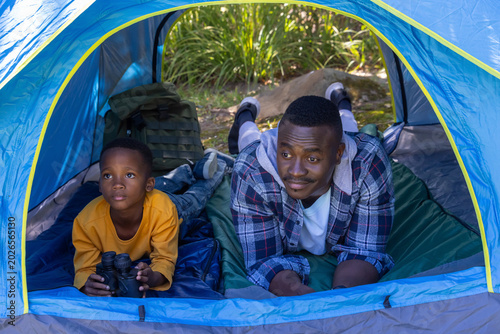 African American father in plaid, son in yellow lying on pads in blue tent holding binoculars