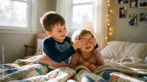 Brother covers his sister's eyes while they play together on their bedroom bed.