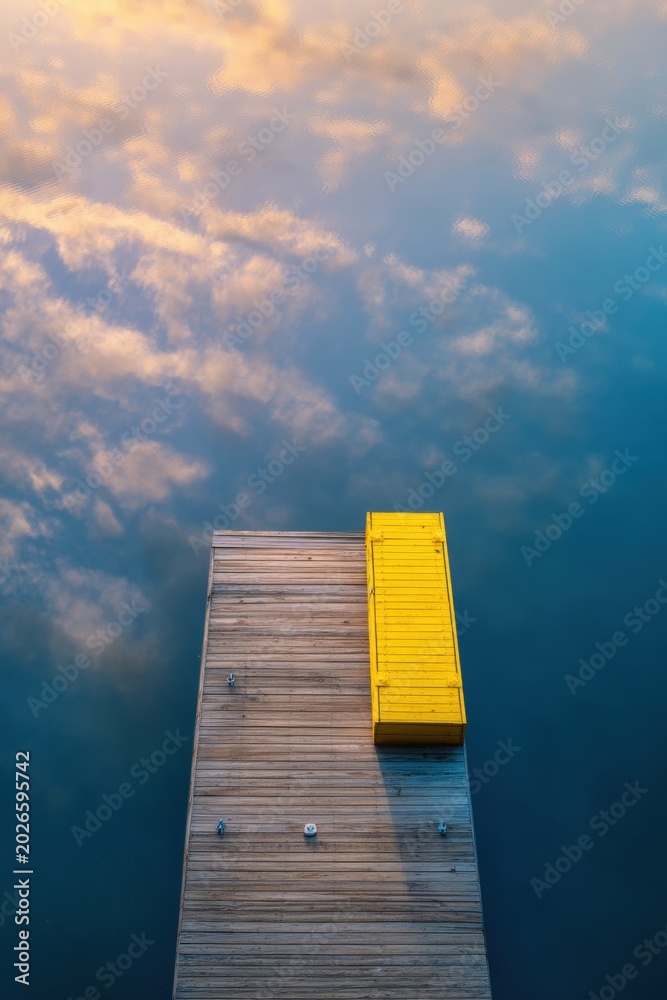 Fototapeta premium Wooden pier with yellow bench on calm lake surface. Aerial view of dock reflecting sky and clouds. Serene nature landscape for vacation, travel meditation and peaceful rest concept.