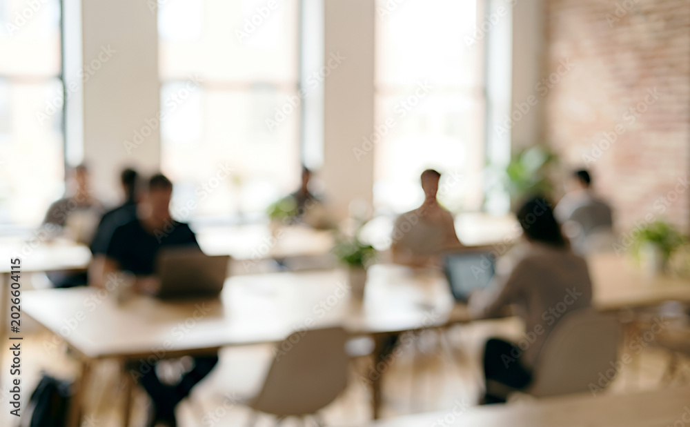 Obraz premium A blurred image of a modern coworking space featuring several individuals working on laptops amidst a bright, open atmosphere with plants.