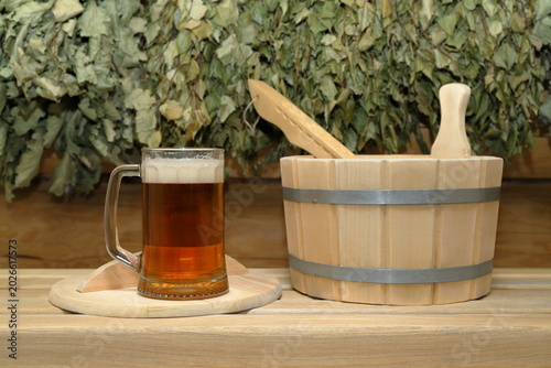 Foto A glass mug of light beer and a wooden vat with a wooden ladle are on a plank bench on the background of a log wall with hanging on it a set of a dry birch and oak brooms