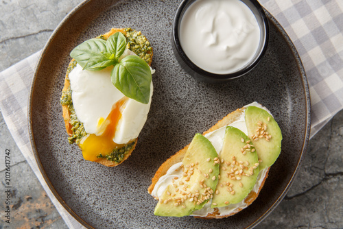 healthy snack of toast with a poached egg and basil pesto, along with avocado and cream cheese, served with sour cream, close-up on a plate on a table. Horizontal top view from above