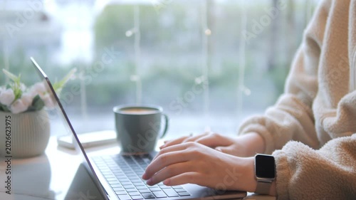 Person types on laptop near window with coffee cup. Hands visible, wearing watch. Background has soft natural light and blurred greenery. Small potted plant beside laptop