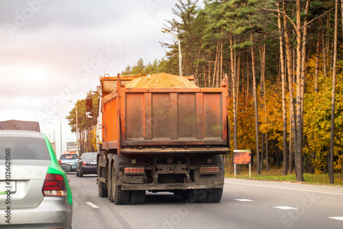 Cuadro en lienzo Rear view of orange dump truck carrying sand on highway with other vehicles in traffic, autumn forest lining side of road under overcast sky, transportation of construction materials