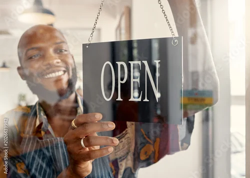 Fototapeta Happy, man and window in restaurant with open sign, small business owner or ready for customer service. Black person, smile and entrepreneur in cafe with welcome board, pride and glass door entrance.