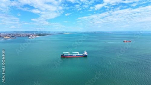 Aerial view of cargo bulk carrier ship in turquoise sea. High angle drone shot of a large blue and red bulk carrier vessel anchored near a coastal port city under a cloudy sky.