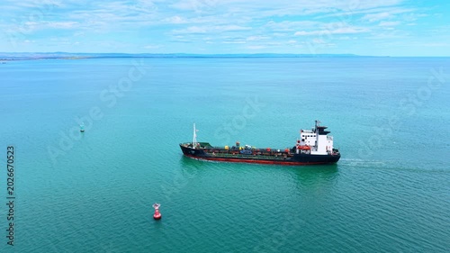 Aerial view of industrial tanker ship sailing in turquoise ocean. Drone view of industrial tanker ship navigating blue sea with red and green navigation buoys under cloudy sky.