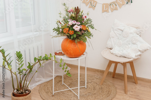 Flower bouquet in carved pumpkin vase on white table. Autumn floral arrangement for Thanksgiving home decoration. Creative floristics design with seasonal plant and orange squash.