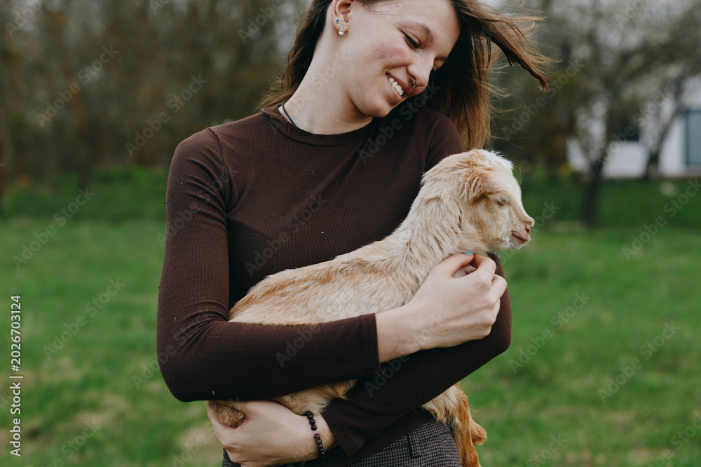 Fototapeta premium Joyful woman outdoors holds a young goat kid in her arms, smiling warmly as they share a tender moment in a green park, showcasing countryside lifestyle and a gentle bond.
