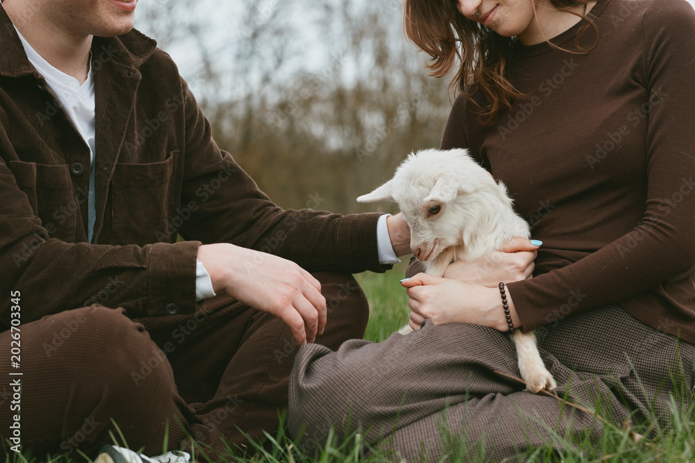 Fototapeta premium Lifestyle outdoor scene shows a couple sitting on grass, gently holding a goat kid, a calm interaction with animal and nature, warm smiles, casual clothing, peaceful family vibe and connection