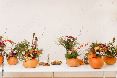 Florist arranging various flower bouquets inside hollowed pumpkins on white table. Autumn floral decor for interior and festive thanksgiving party holiday composition art.