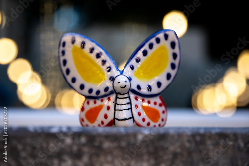 white butterfly, made of glazed ceramic, colored with yellow, orange, and dark blue, with a background of lights, standing on a stone base