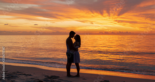 Couple standing on the beach at sunset. Man and woman embracing and looking at each other by the ocean. Romantic vacation and travel concept. Relationship and love during summer holiday.