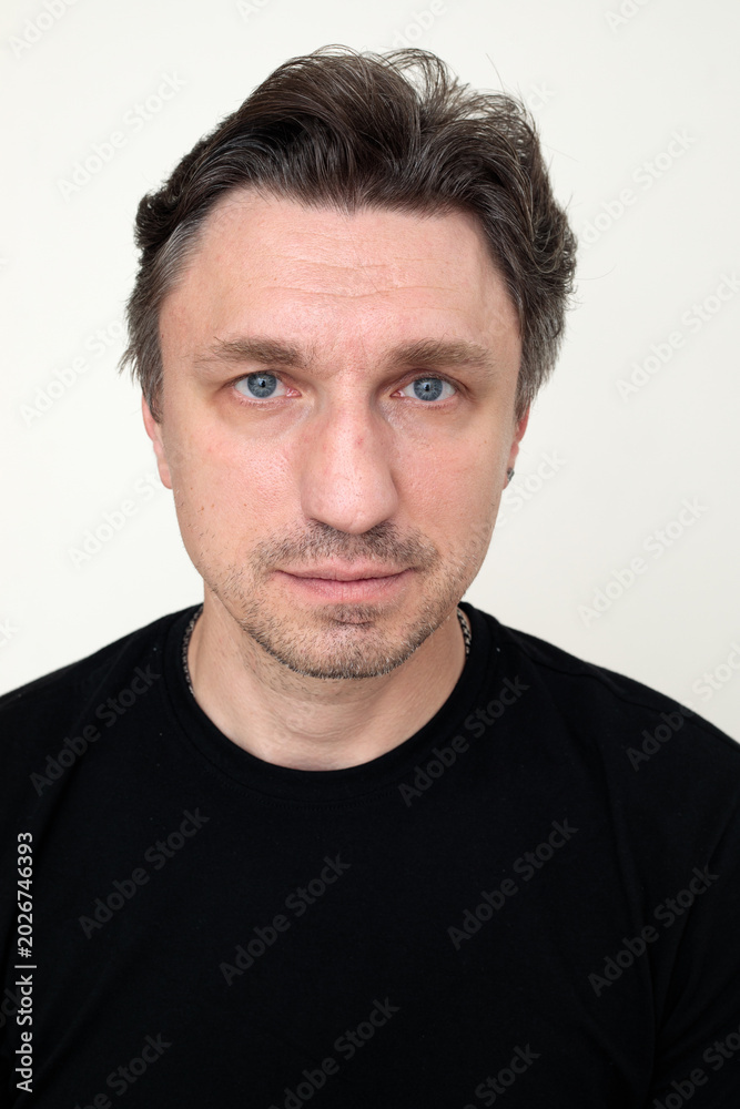 Fototapeta premium close up portrait of serious middle aged man with blue eyes and stubble in black t-shirt looking directly at camera, white background headshot