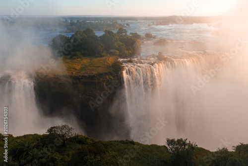 Bild auf Leinwand Aerial sunset view of Victoria Falls, Zimbabwe, shows Zambezi River plunging over basalt cliff, mist rising, islands and channels at the brink, and gorges veiled by spray