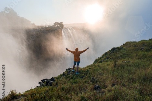 Obraz na plátně An adventurer in an orange jacket stands on a grassy cliff, arms outstretched toward Victoria Falls horseshoe edge, thunderous spray and warm sunset haze on the Zimbabwe side