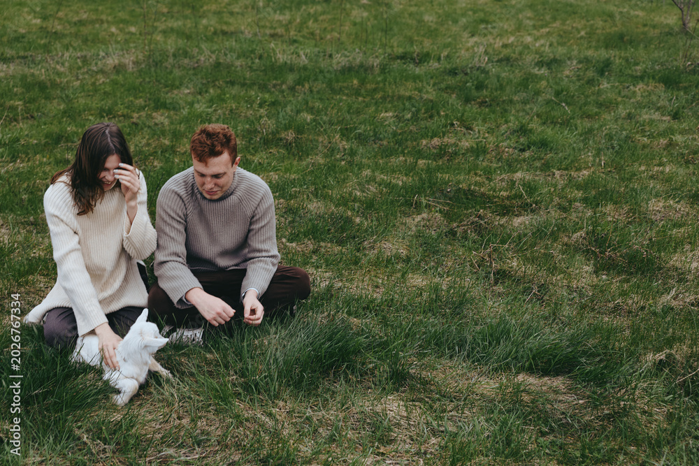 Fototapeta premium Couple sits on grass outdoors sharing a playful moment with a goat kid nearby, creating a warm lifestyle scene focused on companionship and gentle animal interaction in soft light. Cozy.