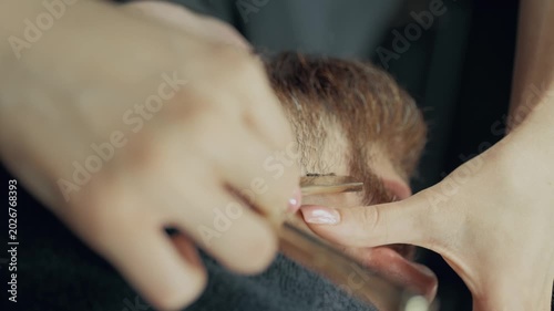 Professional female barber shaving neck of bearded male client with straight razor during grooming procedure in modern barbershop