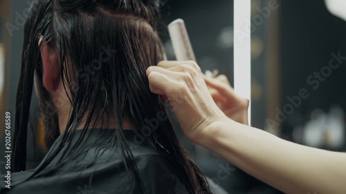 Female barber cutting long wet hair of male client with scissors and comb during professional haircut procedure in modern barbershop