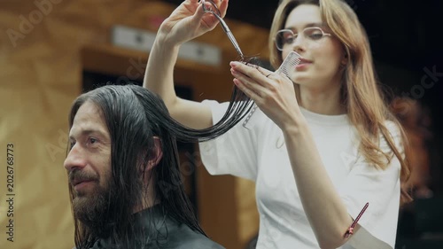 Female barber cutting long wet hair of male client with scissors and comb during professional haircut procedure in modern barbershop