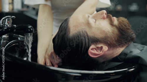 Female barber washing hair of bearded man at salon sink during professional grooming and hair care procedure in modern barbershop